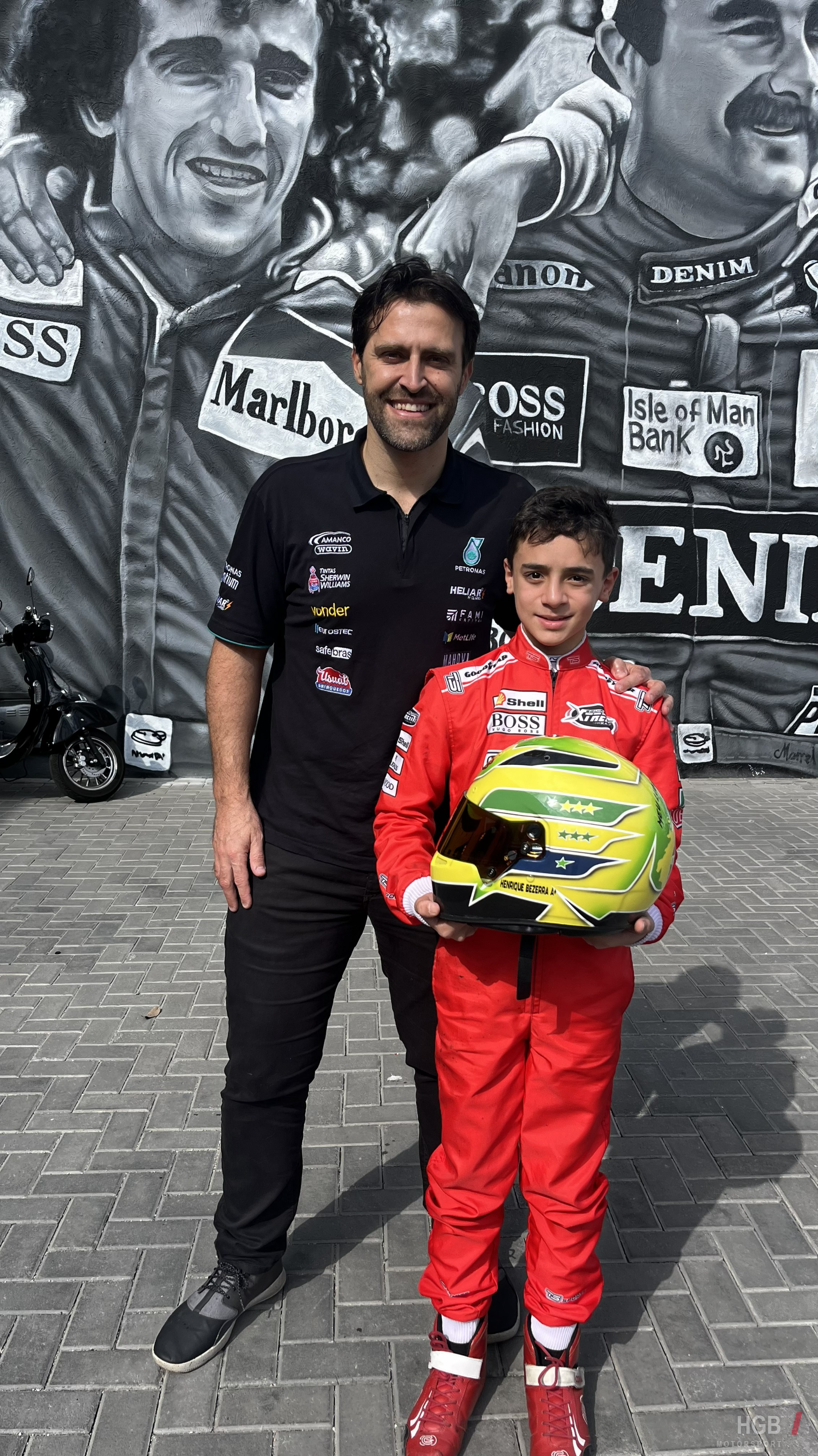 Henrique ao lado do piloto Átila Abreu, fundador da Scuderia Bandeiras, em frente a um mural de Ayrton Senna